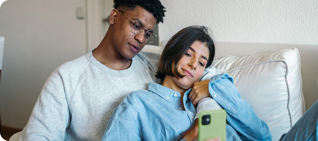 Ein Paar sitzt gemütlich auf einem Sofa, die Frau lehnt sich an den Mann und beide schauen auf ein Smartphone.