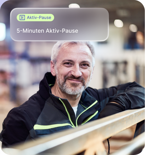 Ein lächelnder Mann mit grauen Haaren und Bart lehnt sich auf ein Geländer in einer Werkstatt oder Fabrikumgebung.