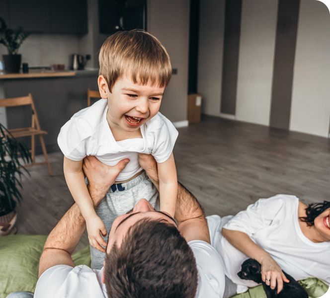 Vater hält lachenden kleinen Jungen in die Luft, während eine Frau mit schwarzem Hund im Hintergrund lächelt.