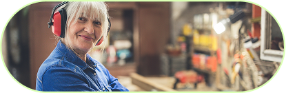 Ältere Frau mit blonden Haaren trägt Schutzkopfhörer und lächelt in einer Werkstatt.