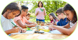 Gruppe von Kindern sitzt an einem Holztisch im Freien und malt mit Pinseln auf Papier, ein Erwachsener steht im Hintergrund.