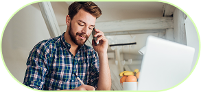 Mann mit kariertem Hemd telefoniert und macht Notizen vor einem Laptop.