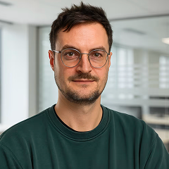 Porträt eines Mannes mit braunen Haaren und Brille, der einen dunkelgrünen Pullover trägt und in einem modernen Büro steht.