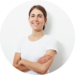 Frau mit braunen Haaren, die in einem weißen T-Shirt vor einer weißen Wand lächelt und die Arme verschränkt hat.