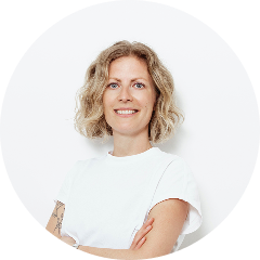 Frau mit blonden, lockigen Haaren in weißem T-Shirt, die lächelt und die Arme verschränkt.