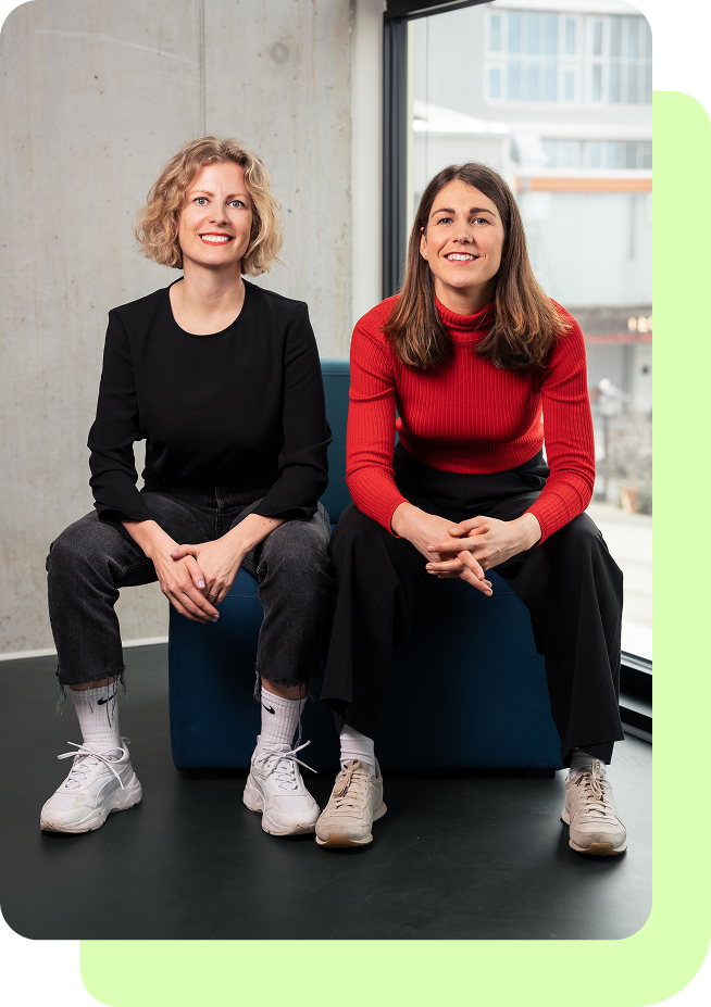 Zwei lächelnde Frauen sitzen nebeneinander auf einem blauen Sitzmöbel vor einem großen Fenster und einem Betonwandhintergrund.