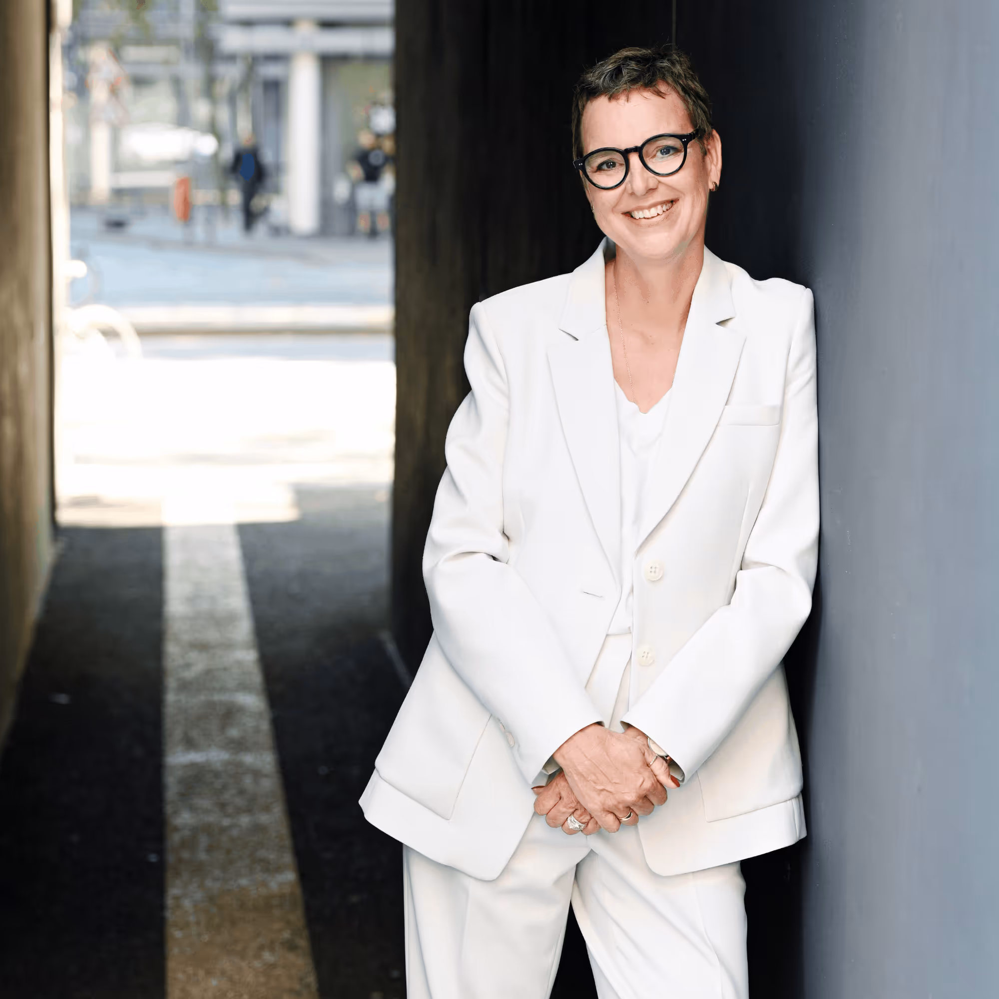 Frau mit kurzen Haaren und Brille, die in einem hellen Anzug lächelnd an einer Wand lehnt.