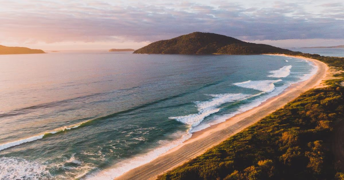 Curved sandy beach with waves and forested hills under a cloudy sky at sunset.