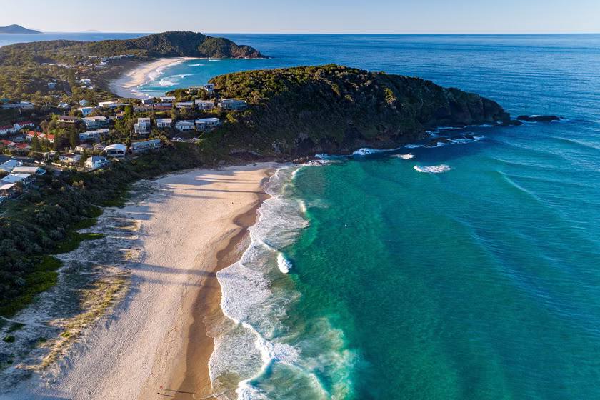 Aerial view of a coastal town with sandy beaches, green hills, and turquoise ocean waves.