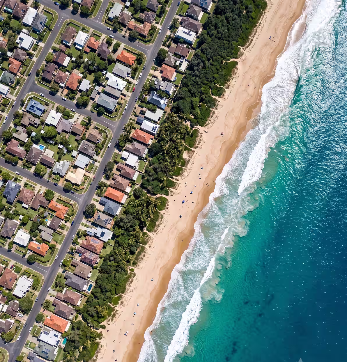 Aerial view of houses and streets alongside a sandy beach and ocean waves with scattered people.