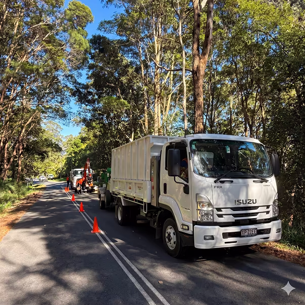 White Isuzu FRR truck parked on a forest-lined road with orange safety cones leading to a vehicle with an excavator attachment.