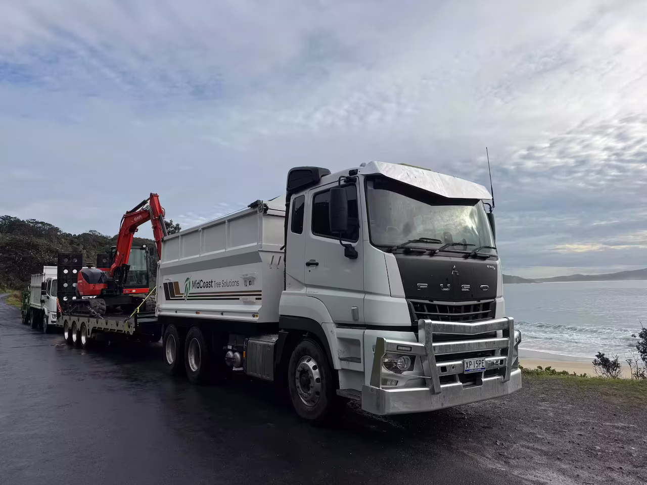 White Fuso truck with trailer carrying red excavator, parked near a coastal shoreline under cloudy sky.