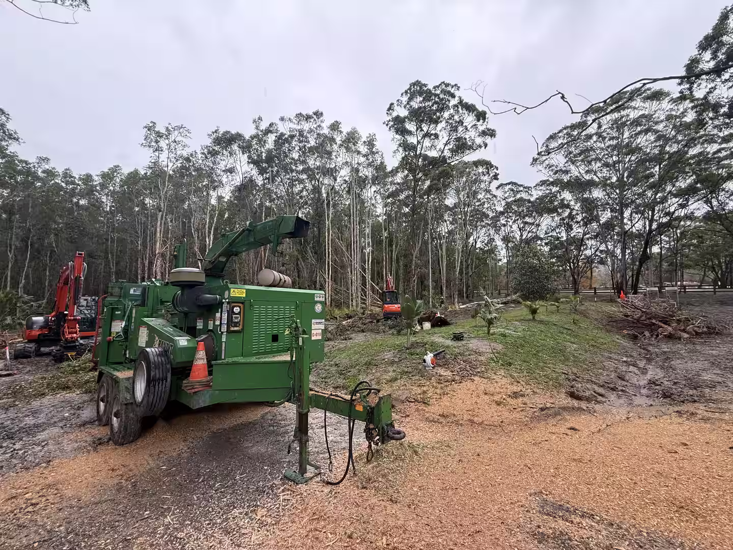 Green wood chipper and red excavators at a forest clearing with wood chips on the ground.