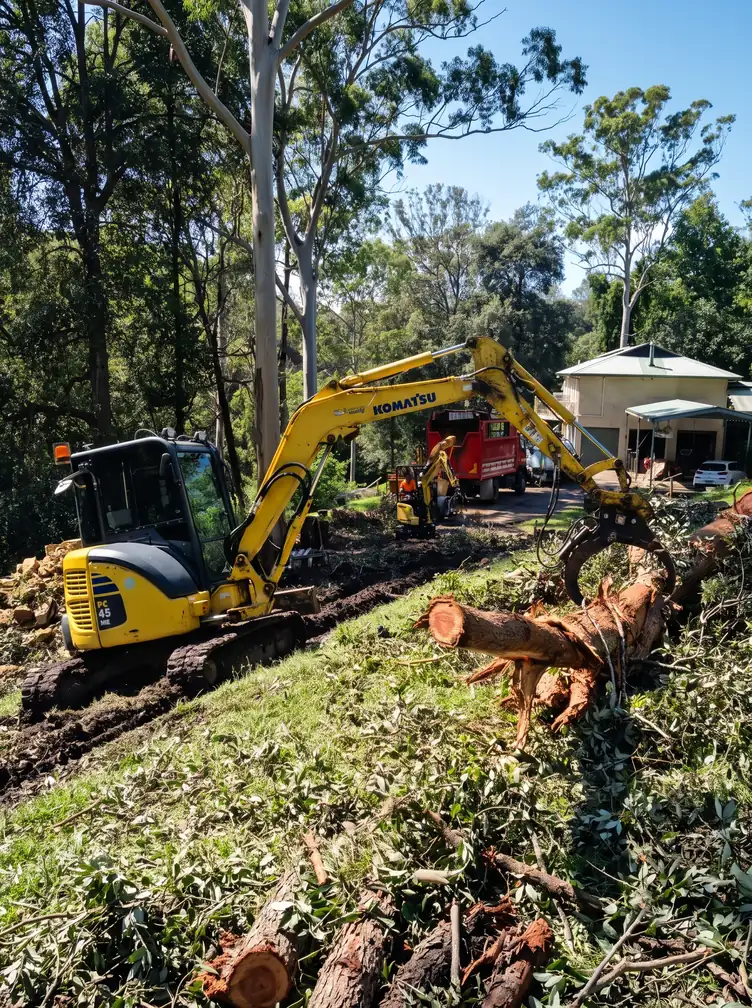 Yellow excavator with a grapple clearing felled trees on a wooded hillside near a house.