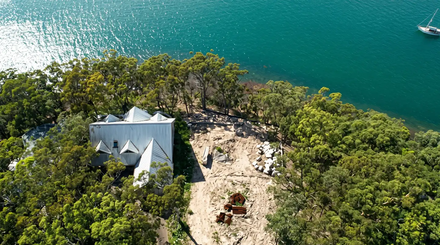 Aerial view of a modern house with a geometric pyramid roof nestled among trees on a sandy waterfront, turquoise water beyond.