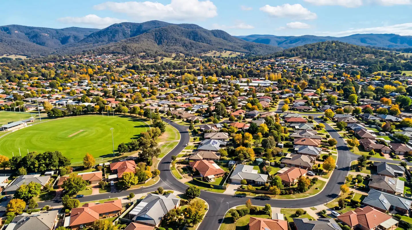 Aerial view of a suburban town with red-roofed houses, a sports oval, and forested mountains in the background.