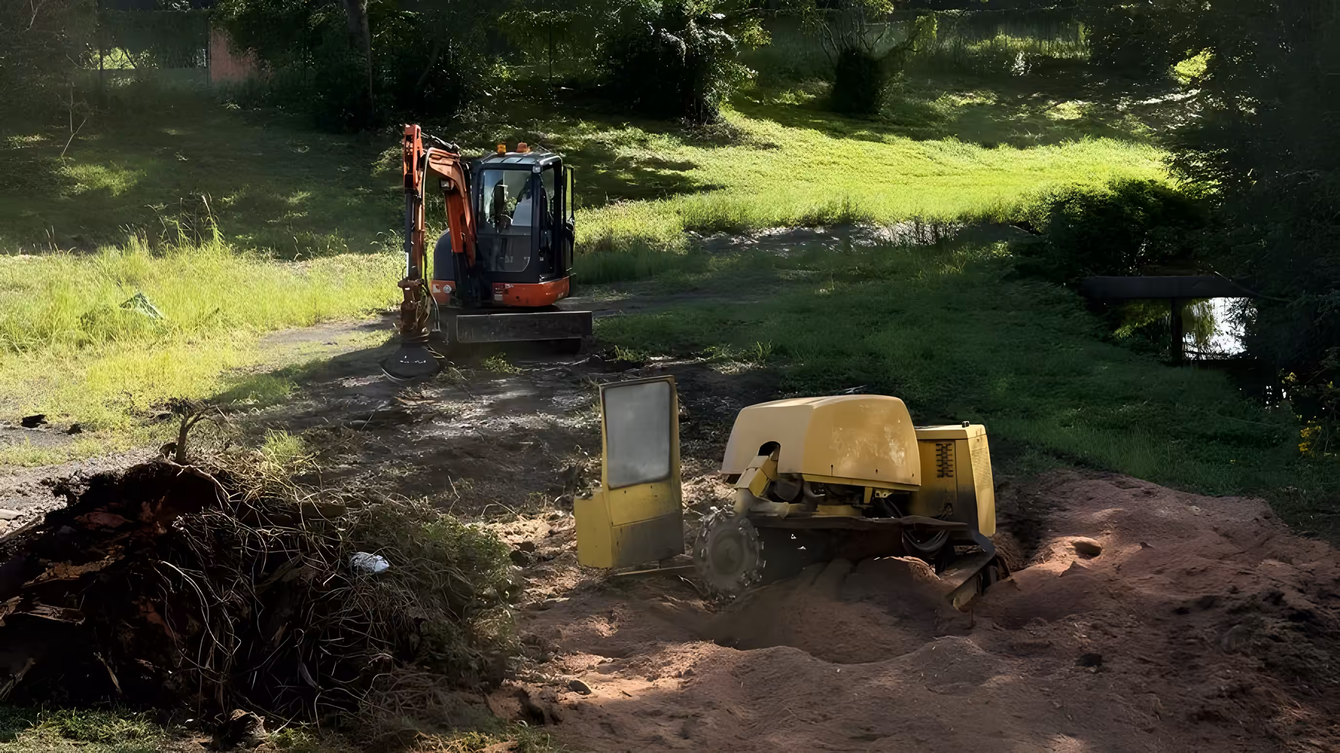 Stump grinder and excavator removing a tree stump and root ball on a grassy rural property.