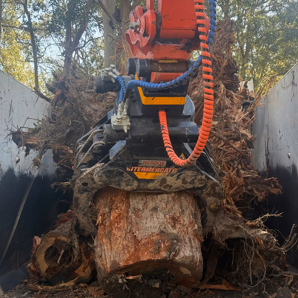 Excavator grapple lifting a large stump and root ball out beside a fence line