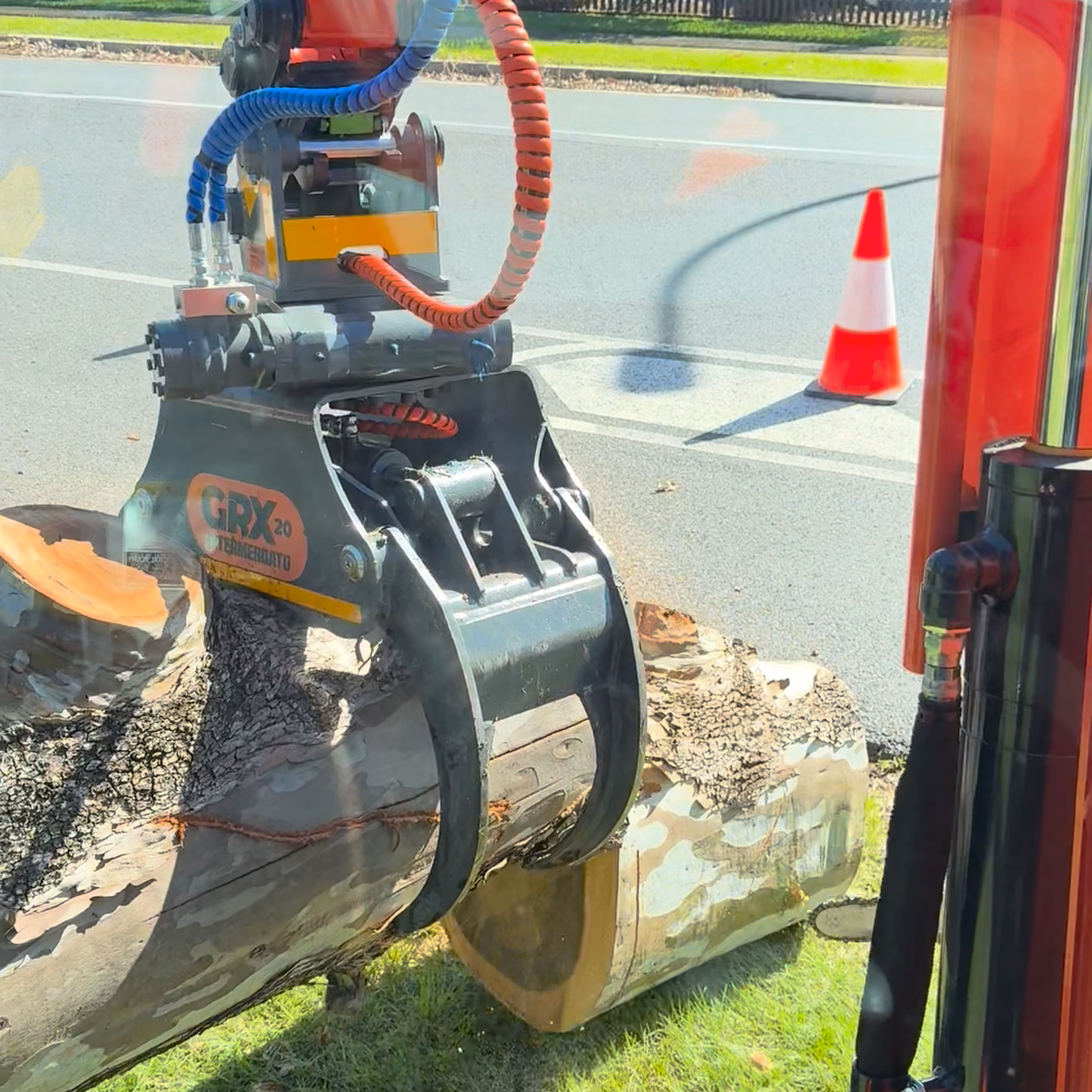 Excavator grapple lifting a fallen log off a roadside verge during storm cleanup