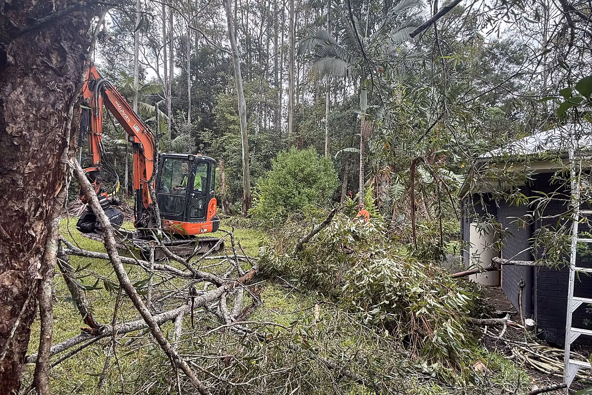 Excavator clearing storm-damaged tree debris near a house