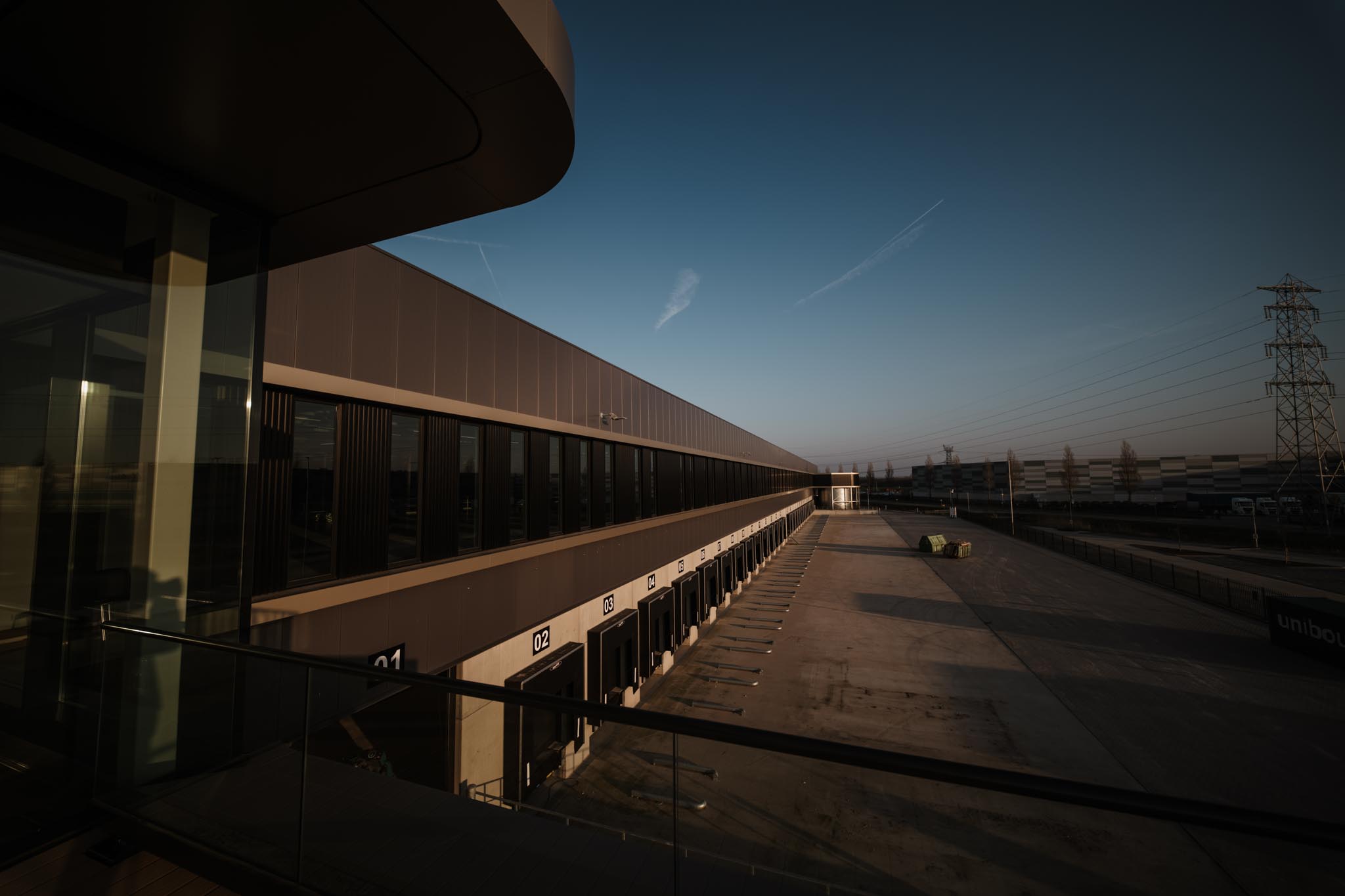 Empty loading dock area of a large industrial warehouse building at dusk with numbered loading bays and a clear sky.
