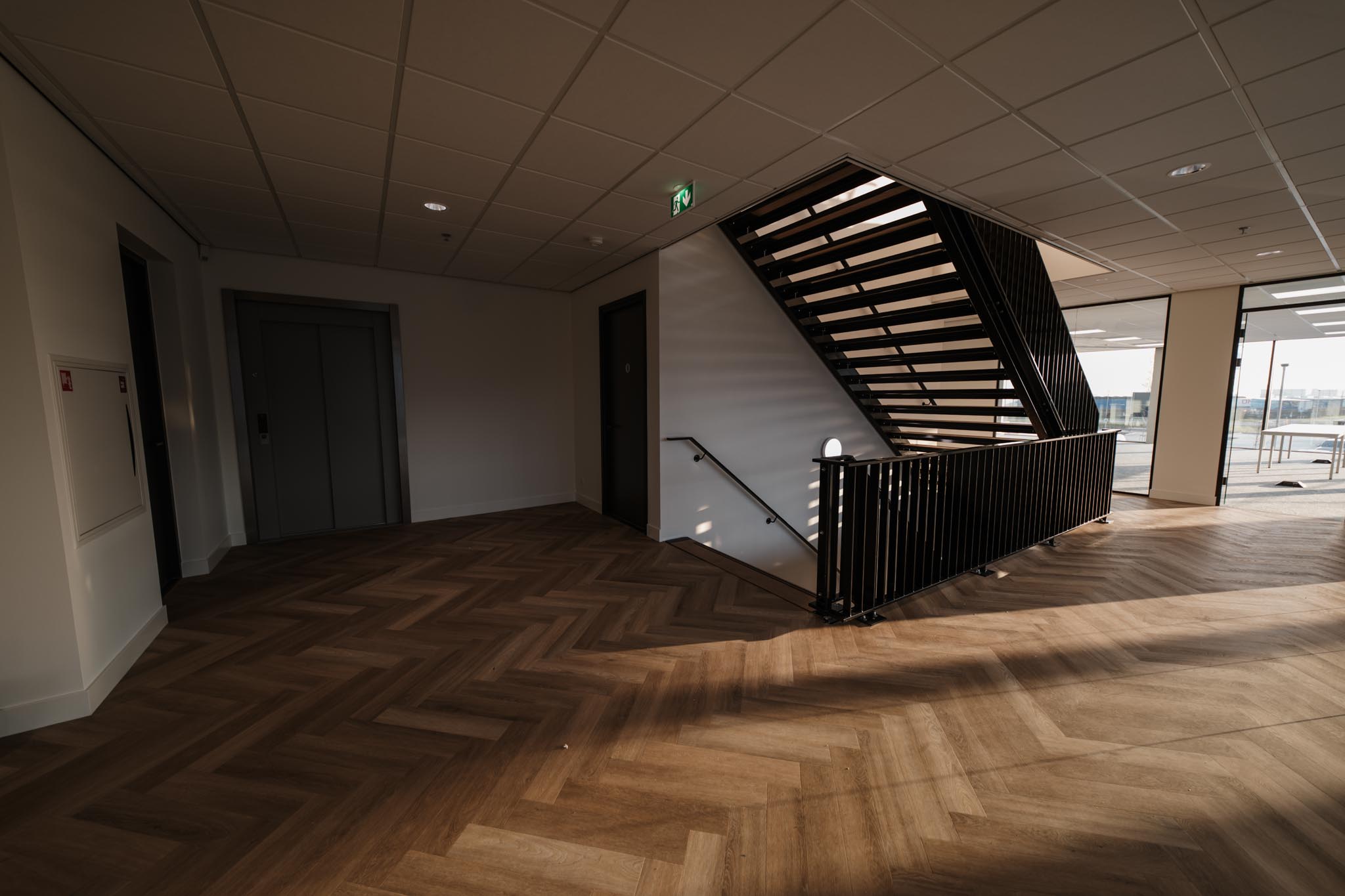 Modern interior hallway with wood-patterned flooring, elevator doors, stairs with black railing, and sunlight streaming through large windows.