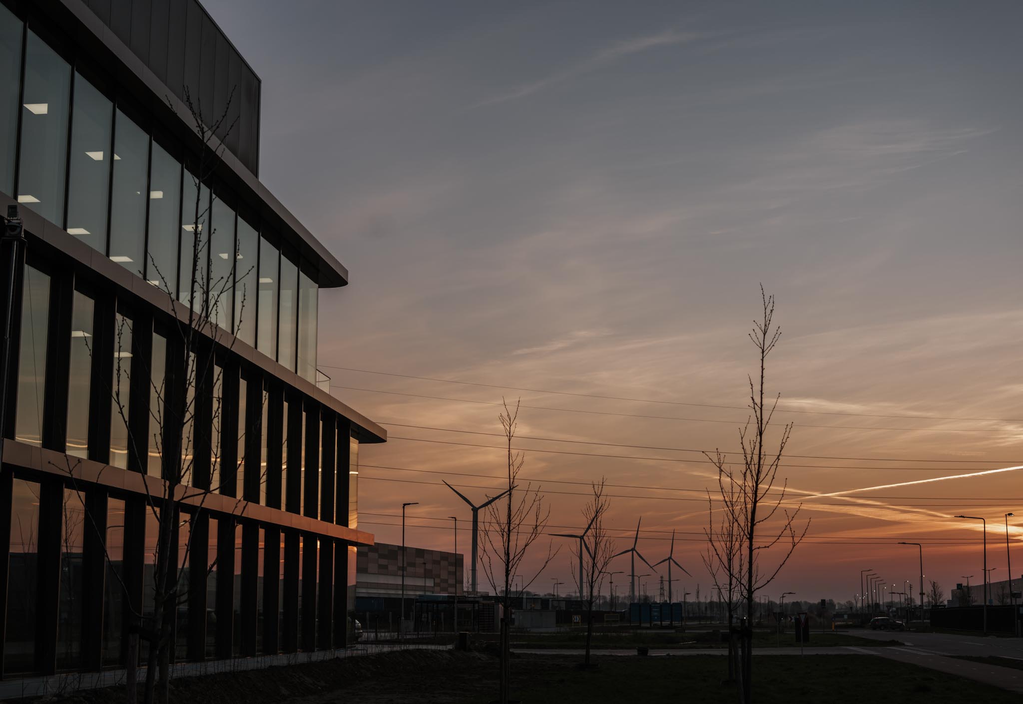Modern glass office building reflecting a sunset with wind turbines and bare trees in the background.