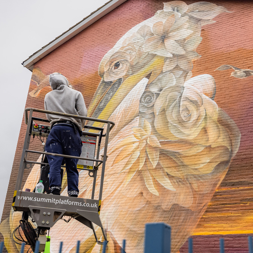 Person on a lift painting a large pelican mural with floral patterns on a brick wall.