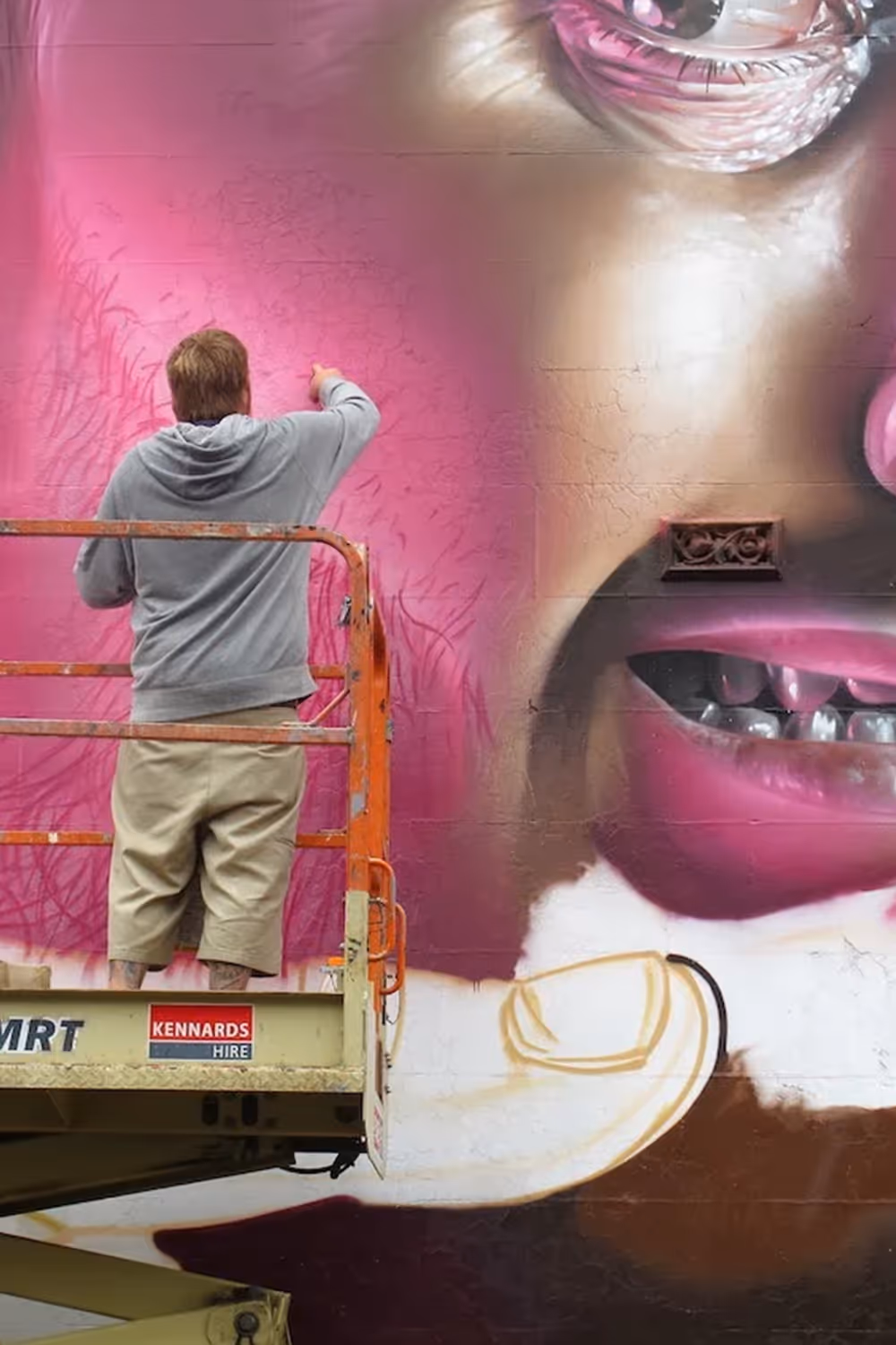 Artist on lift painting a large mural of a face with pink lips on a wall.