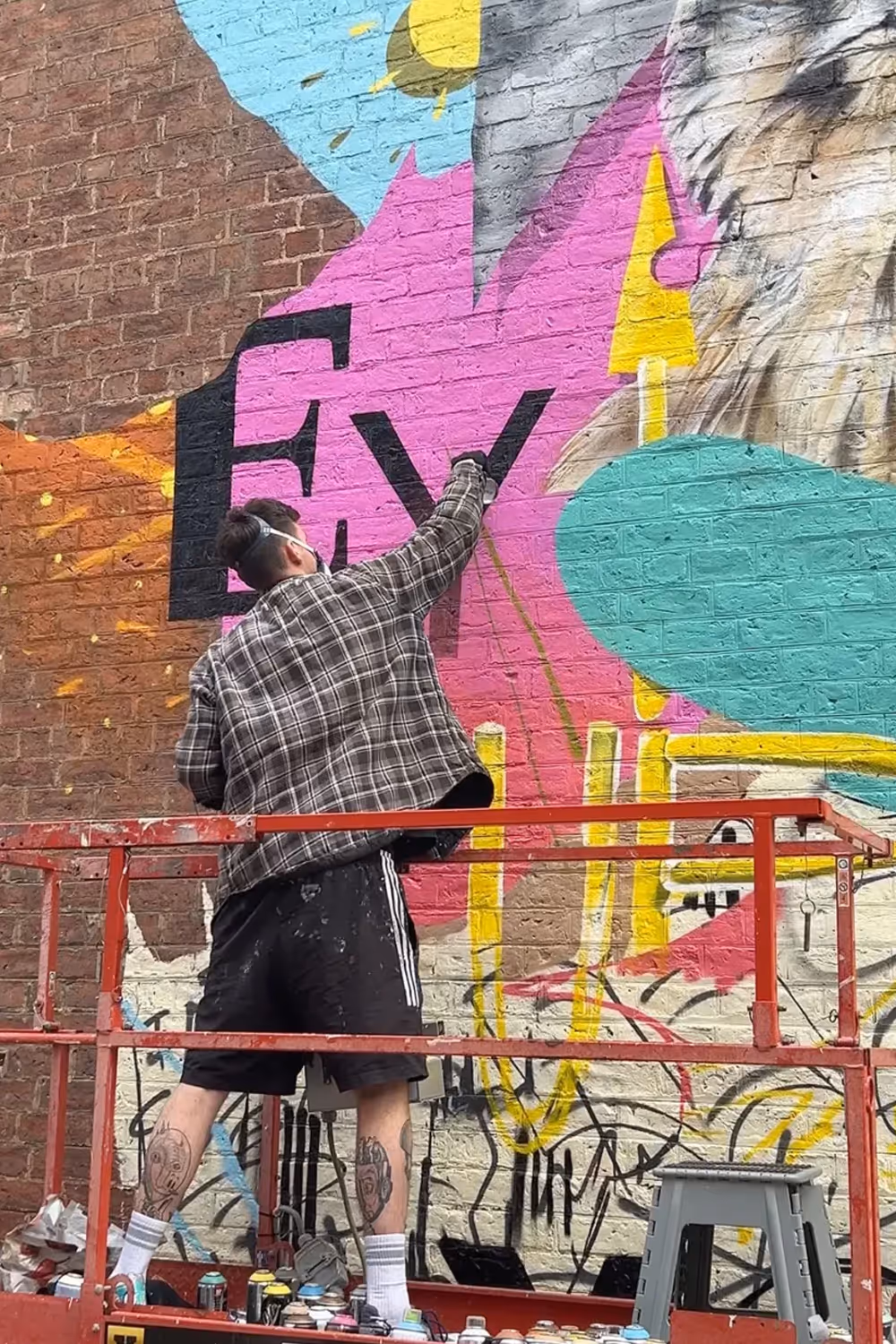 Artist on lift spray-paints colorful mural with large letters and animal face on brick wall.