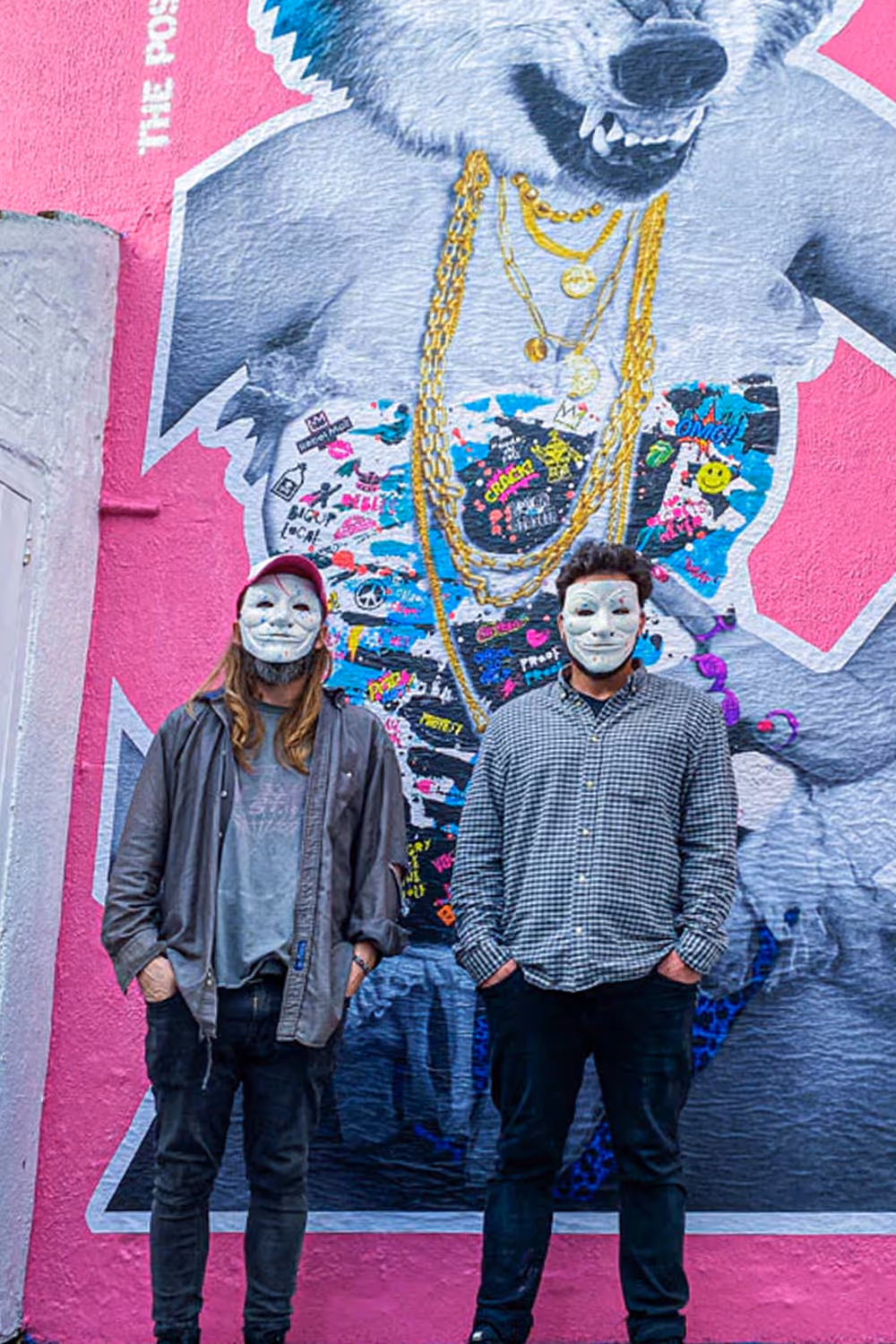 Two men wearing white masks stand in front of colorful graffiti of a wolf with gold chains on a pink wall.
