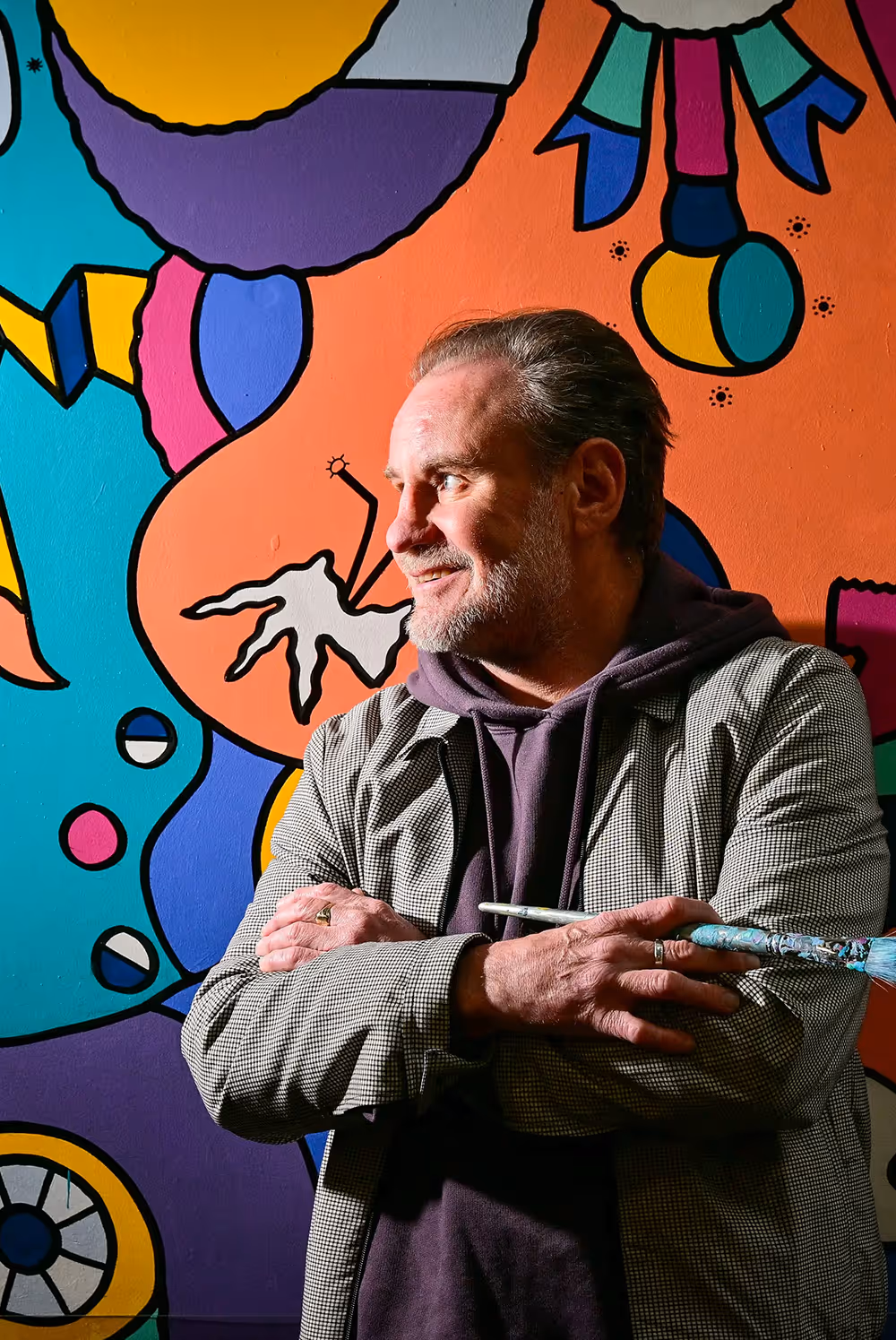 Man holding a paintbrush stands crossed-armed in front of a colorful abstract mural on a wall.