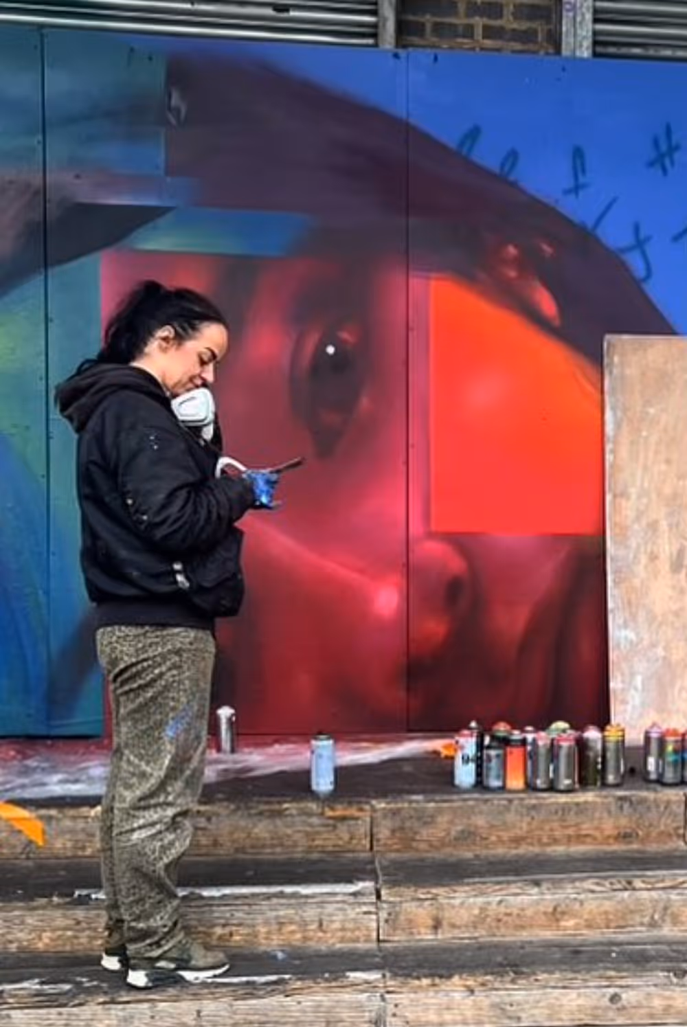 Artist stands with phone and spray paint gloves before large colorful street art mural of a face.
