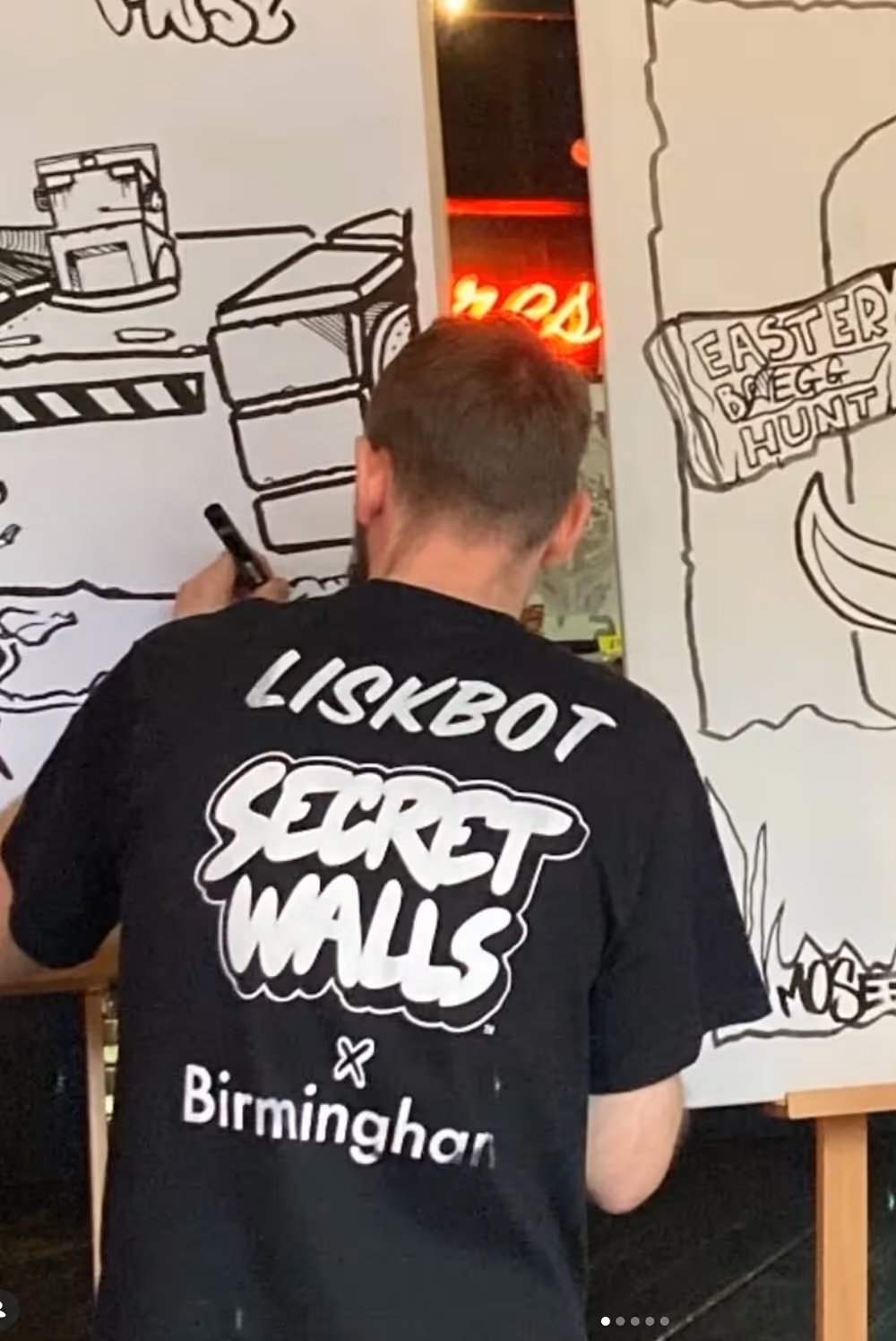 Man wearing a black Liskbot Secret Walls Birmingham shirt drawing cartoon art on white boards.
