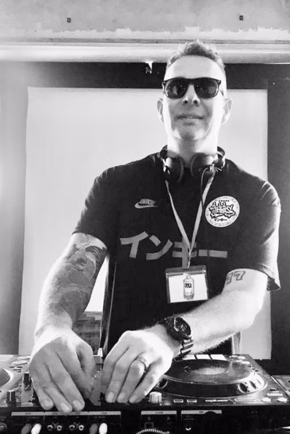 Man with sunglasses and headphones DJing on a mixer, wearing a black shirt with tattoos visible on both arms.