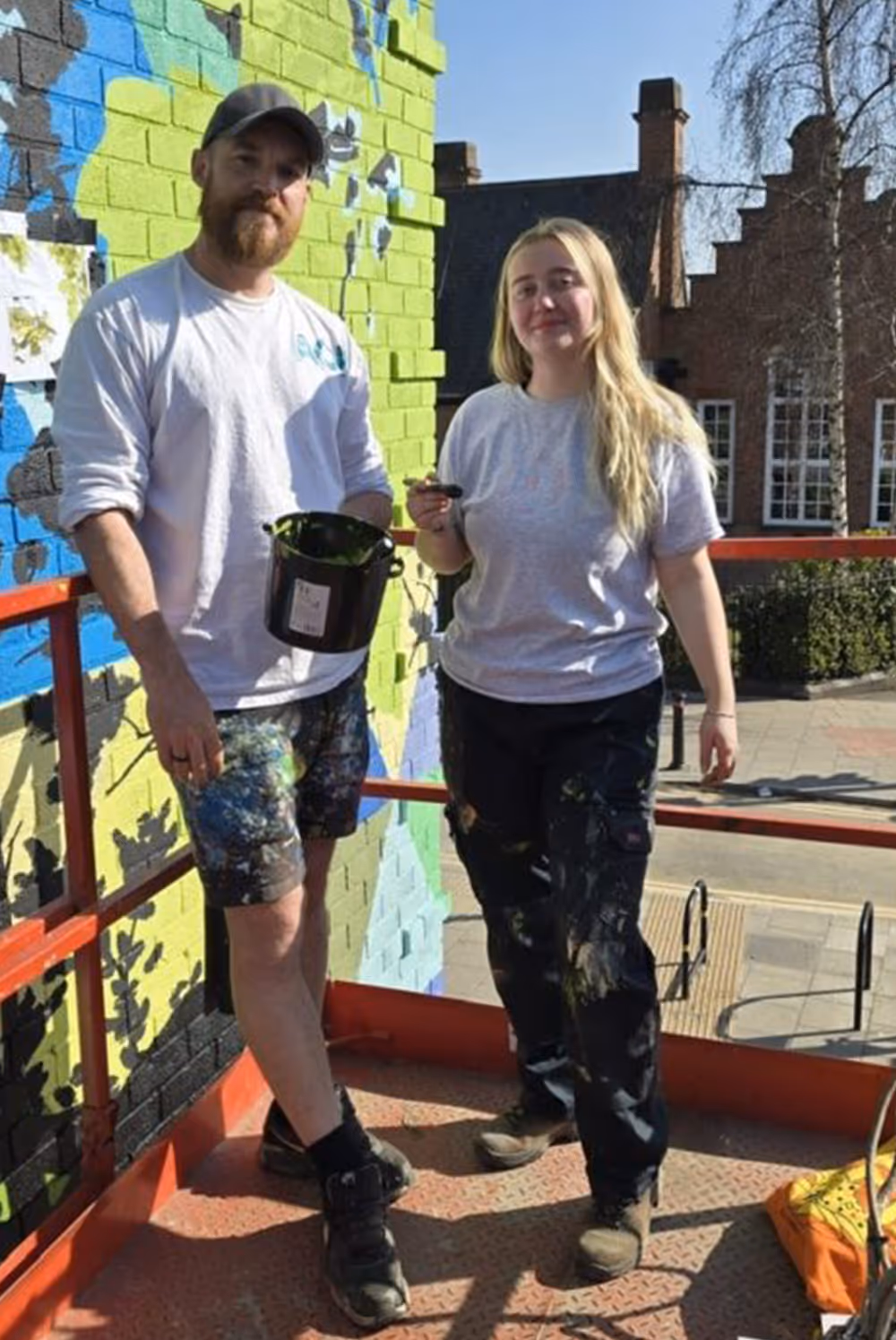 Two painters with paint-splattered clothes standing on a platform next to a brightly painted wall.