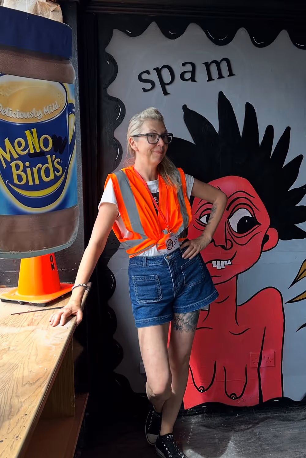 Woman in orange safety vest and shorts standing by a table with mural and large coffee cup behind her.
