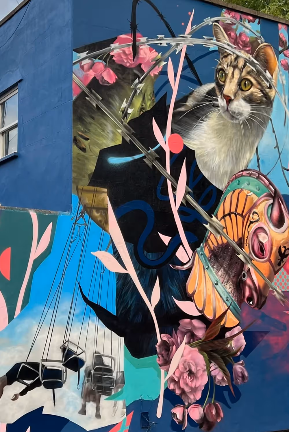 Colorful mural of a cat wearing a collar, flowers, barbed wire, and a swing ride on a blue wall.