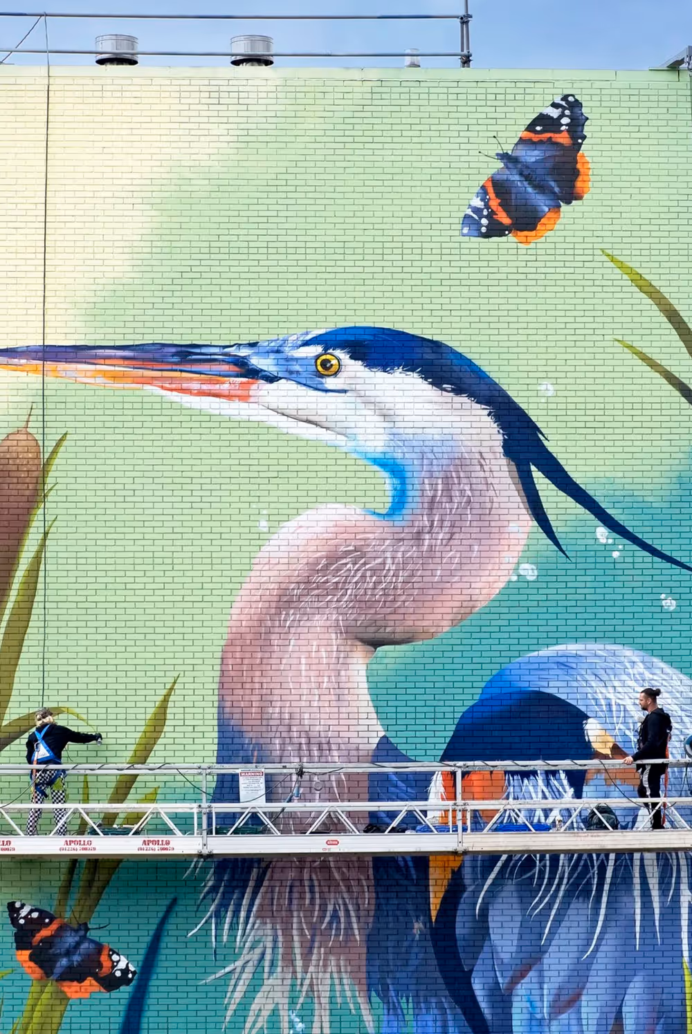 Two workers on a scaffold painting a large mural of a heron and butterflies on a brick wall.