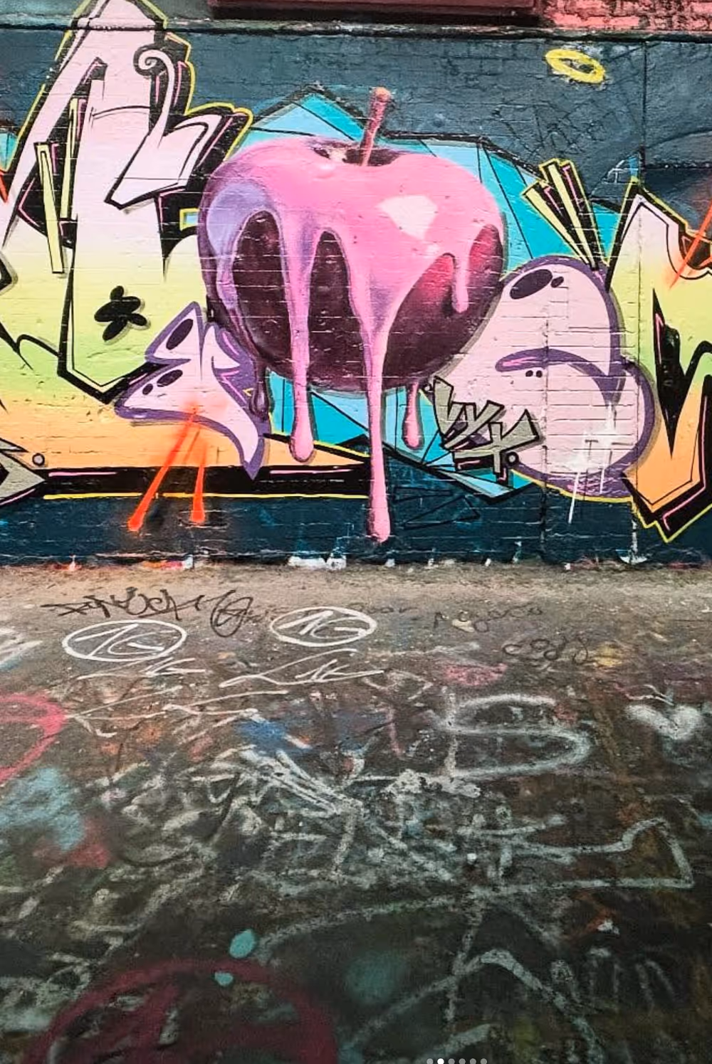Colorful graffiti on a brick wall featuring a large pink apple with dripping paint and abstract shapes.