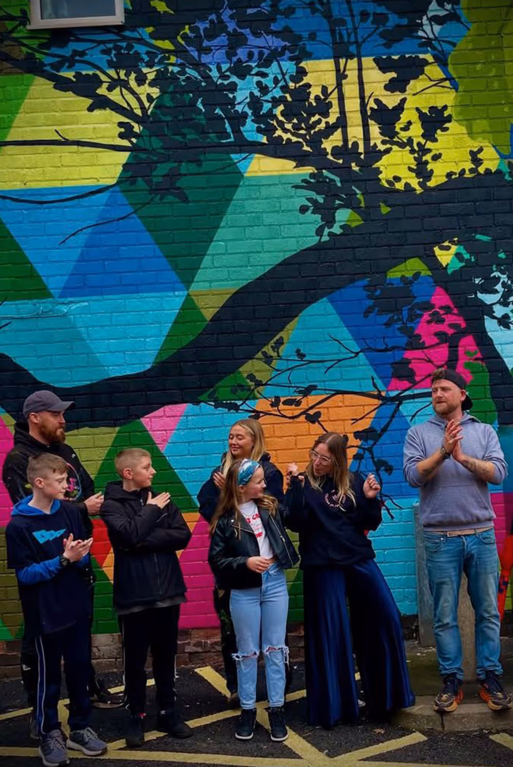 Group of six people standing and clapping in front of a colorful geometric mural with a tree silhouette.
