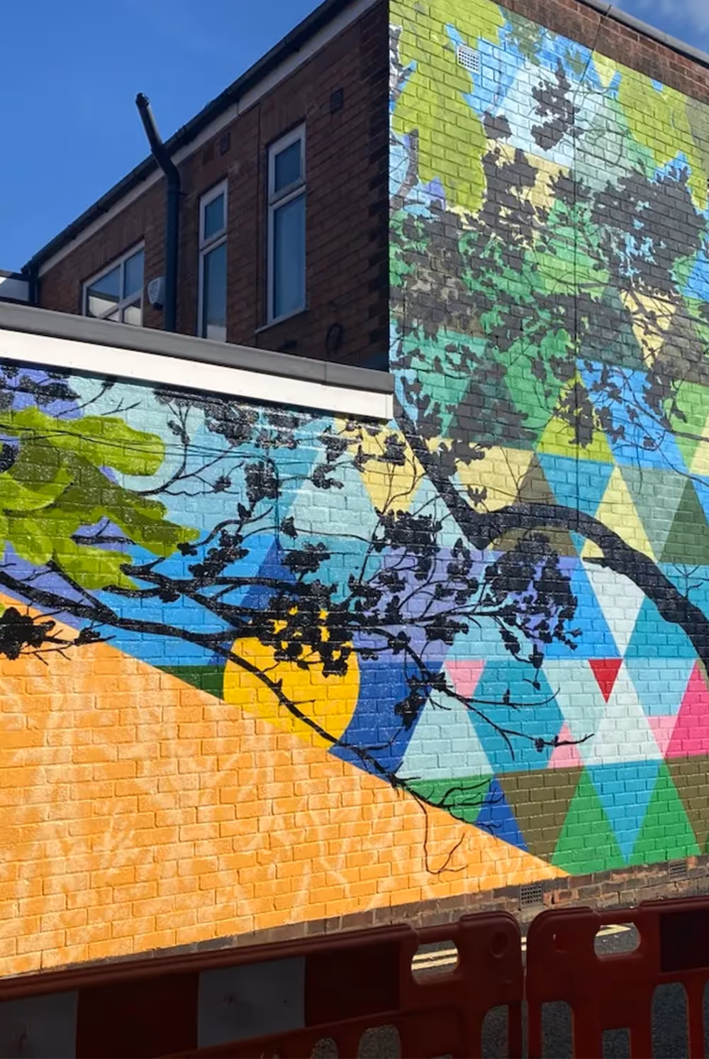 Colorful geometric mural on brick wall with tree branches casting shadows over it.