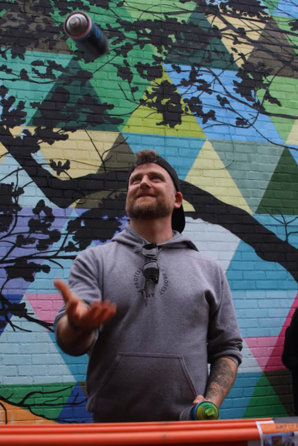 Man in gray hoodie and backwards cap tossing a spray paint can against a colorful mural wall.