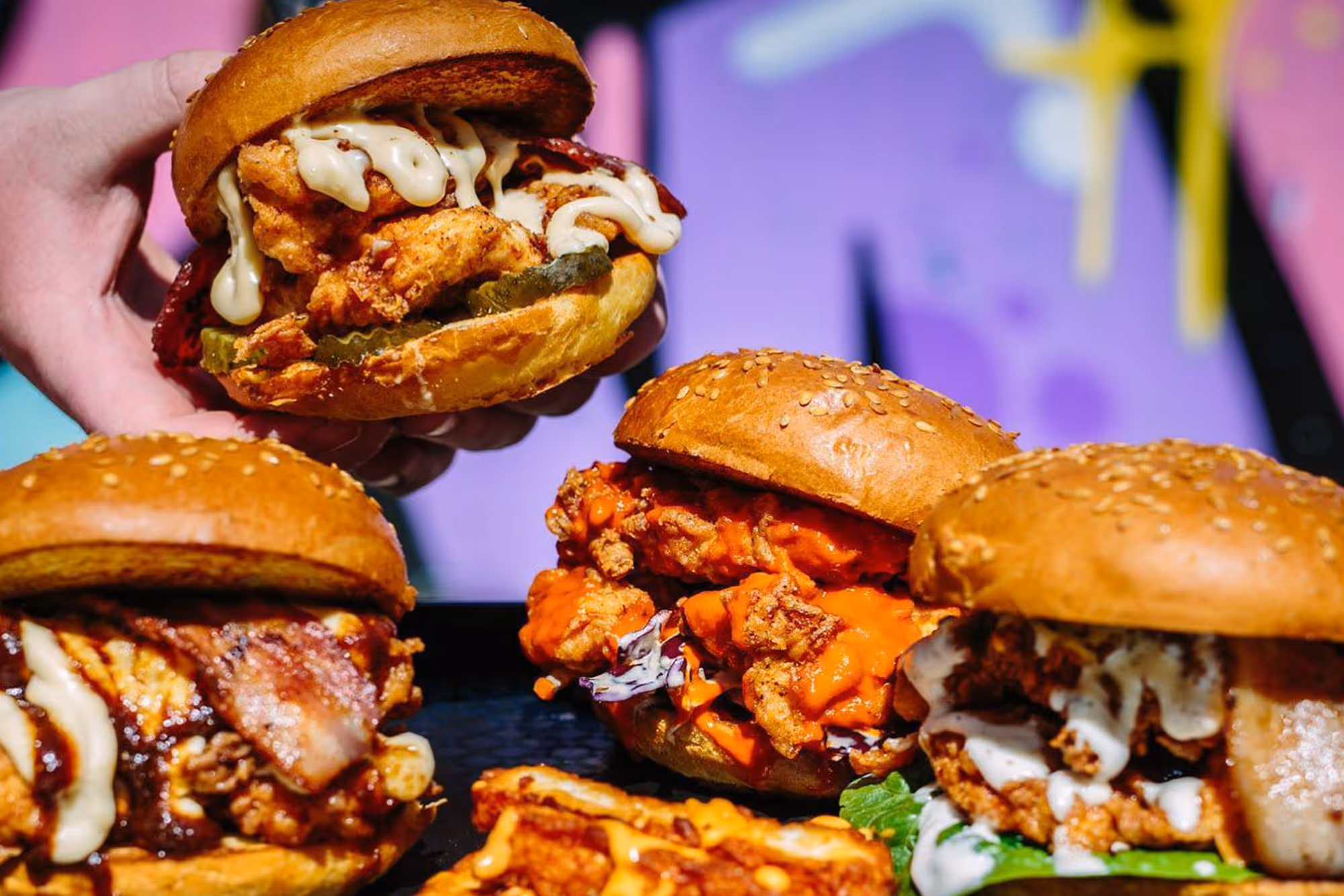 Close-up of four fried chicken sandwiches with various sauces and toppings on sesame buns.