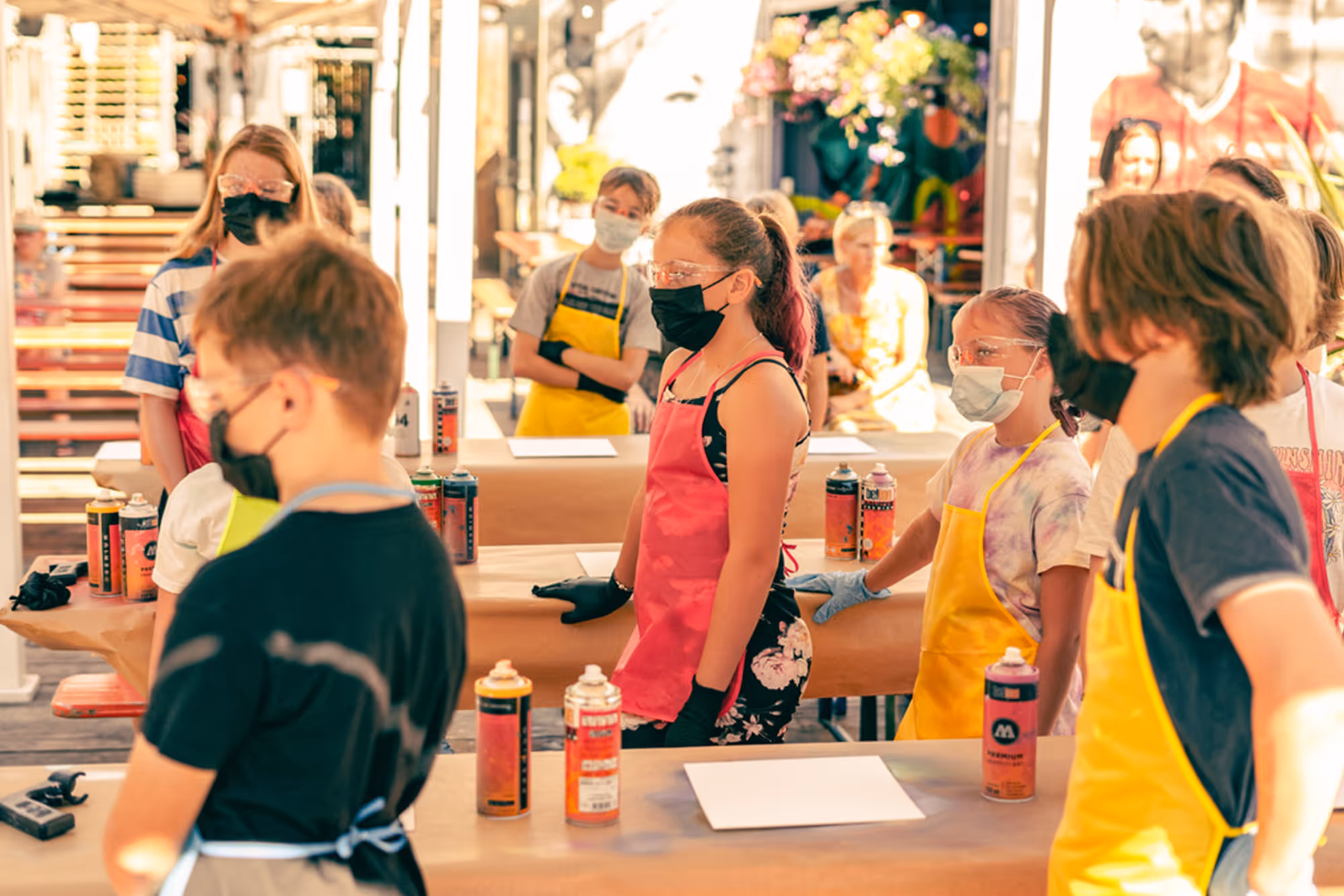 Children wearing aprons and masks standing at tables with spray paint cans for art workshop.