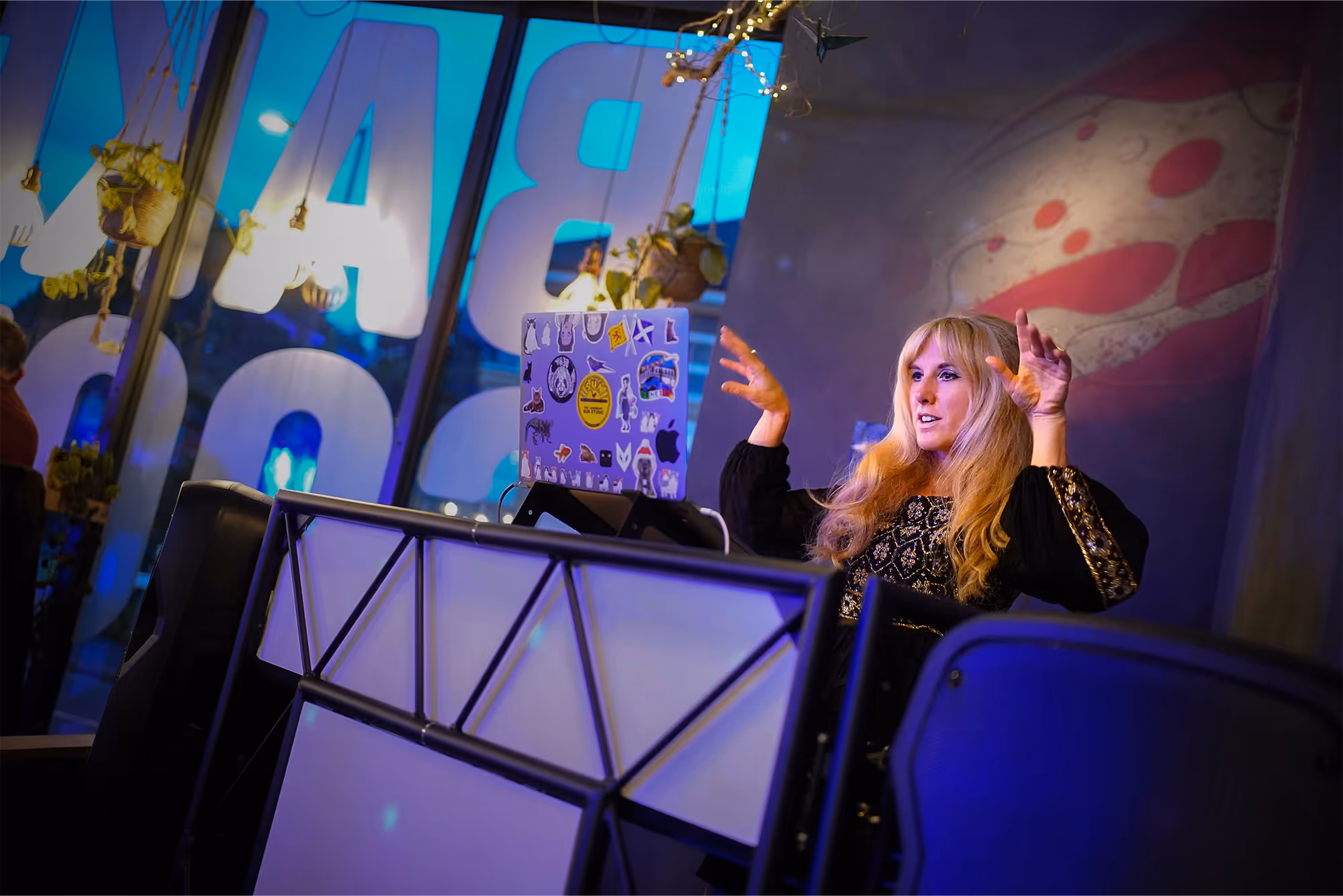 Woman with long blonde hair gestures while sitting behind a decorated laptop in a dimly lit room.