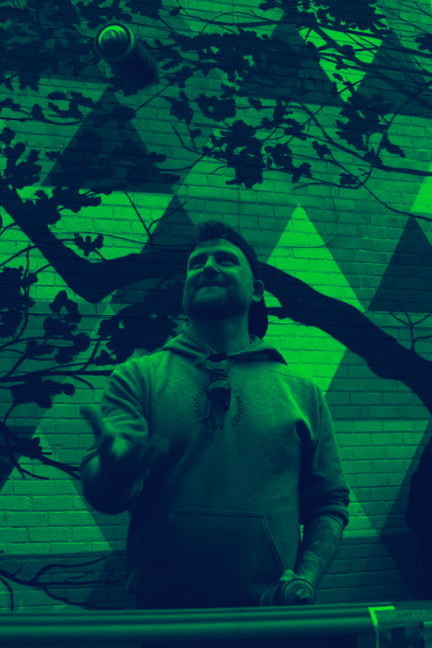 Man wearing a hoodie and backwards cap tossing a spray paint can in front of a graffiti wall.