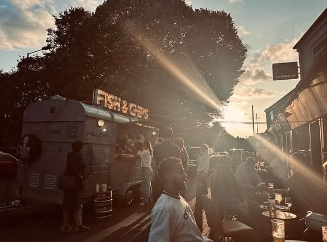 People at an outdoor fish and chips food truck with sunset and tree in the background.
