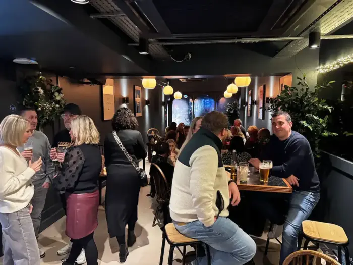 People socializing and drinking beer in a cozy, dimly lit pub with black walls and hanging lights.