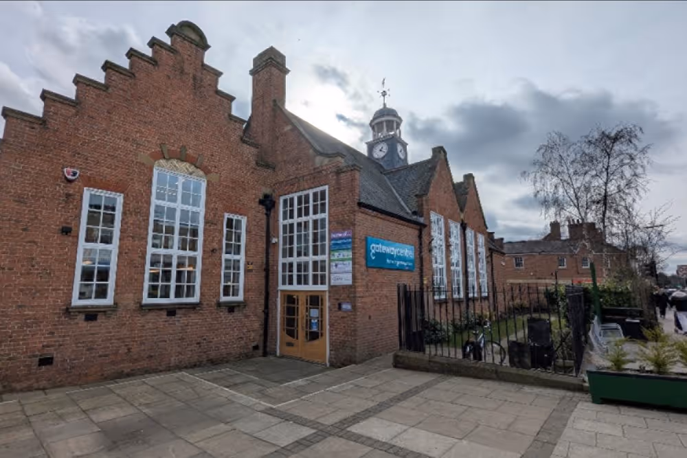 Brick building with large windows, clock tower, and gateway entrance on a cloudy day.